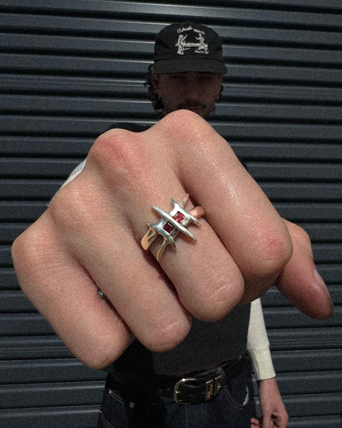 A close-up of a hand wearing a silver ring with a red square cut gemstones, with a man in a cap and jeans standing behind against a dark metal shutter background.