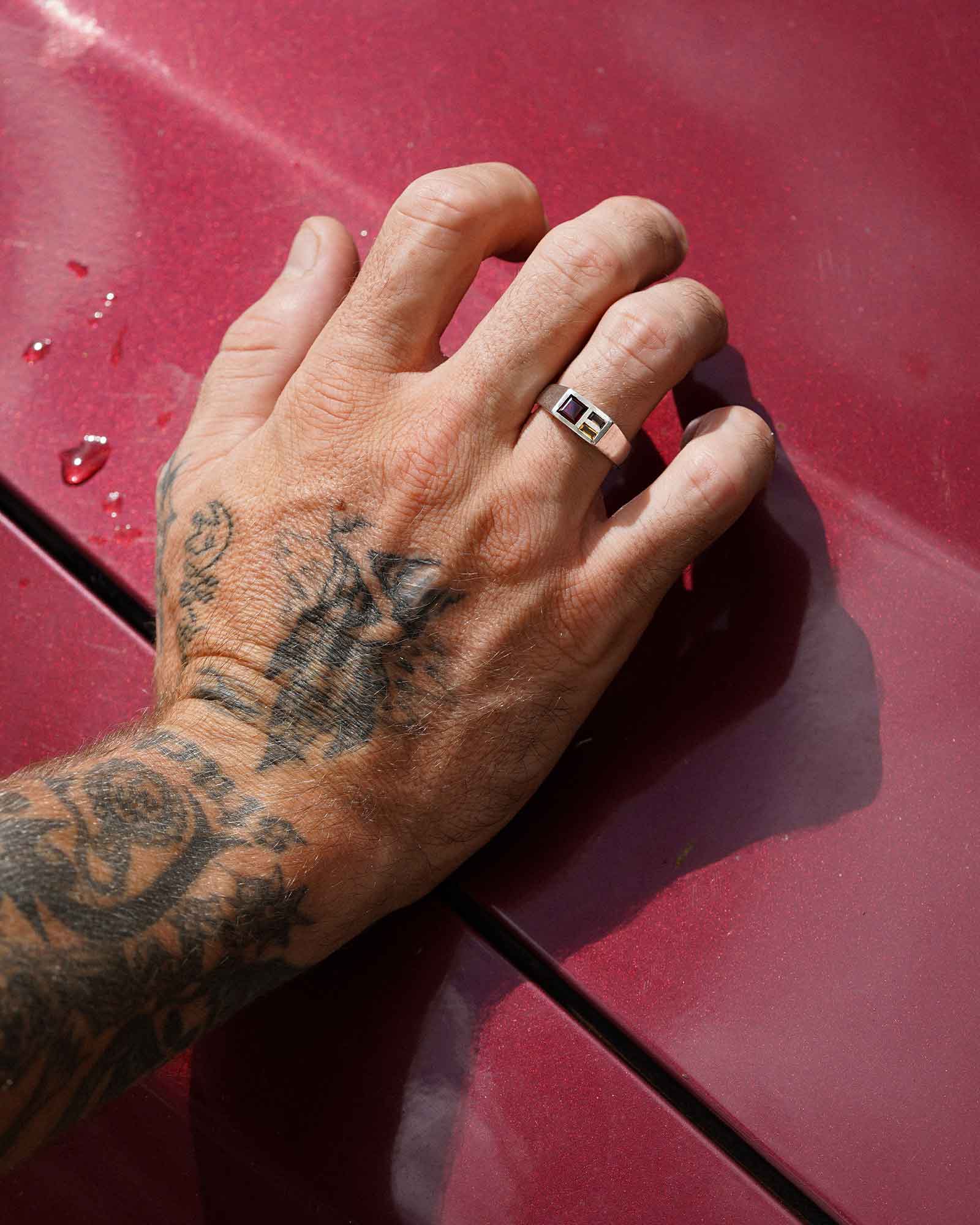 Hand with tattoos on a red car surface wearing a ring with gemstones