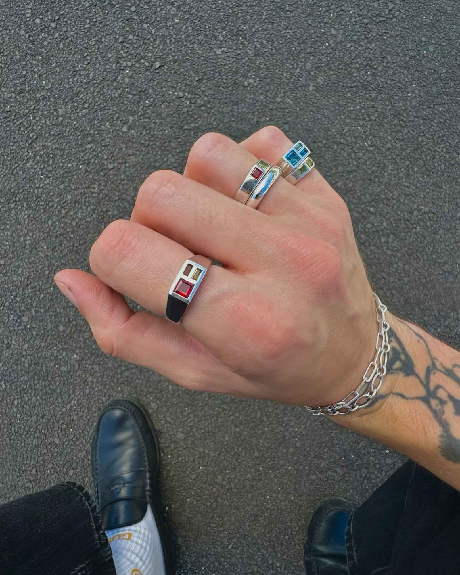 Hand wearing multiple rings with colorful gemstones on a gray pavement background