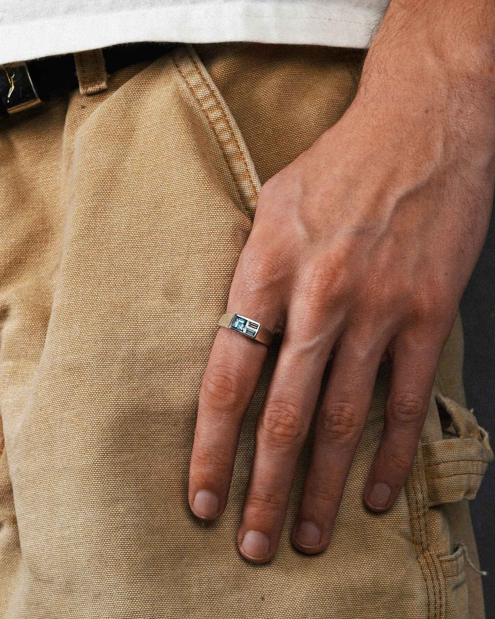Hand wearing a ring with a blue stone on beige carpenter pants.