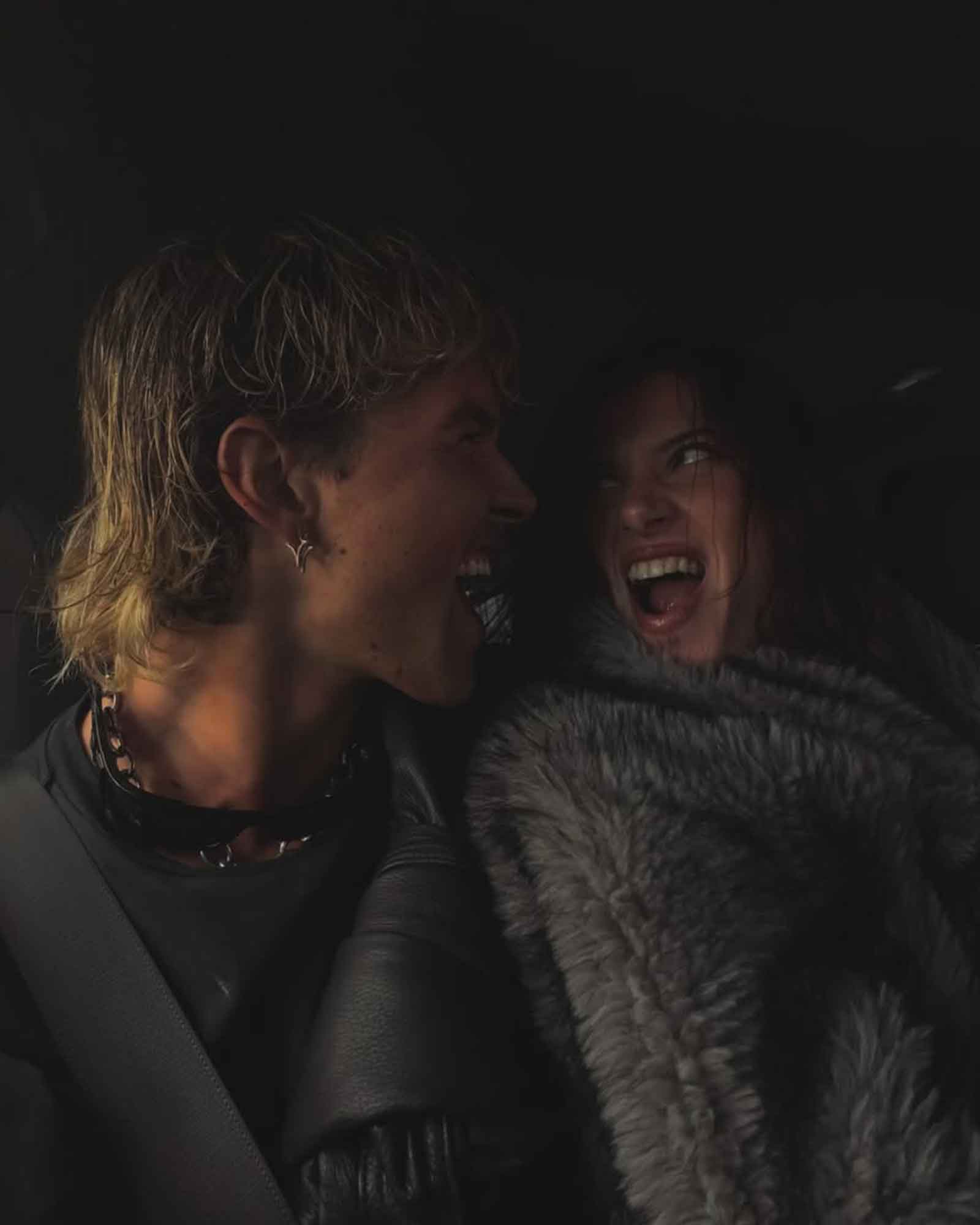 Two people in a dark setting, one with silver star shaped earrings and chains, looking at each other.