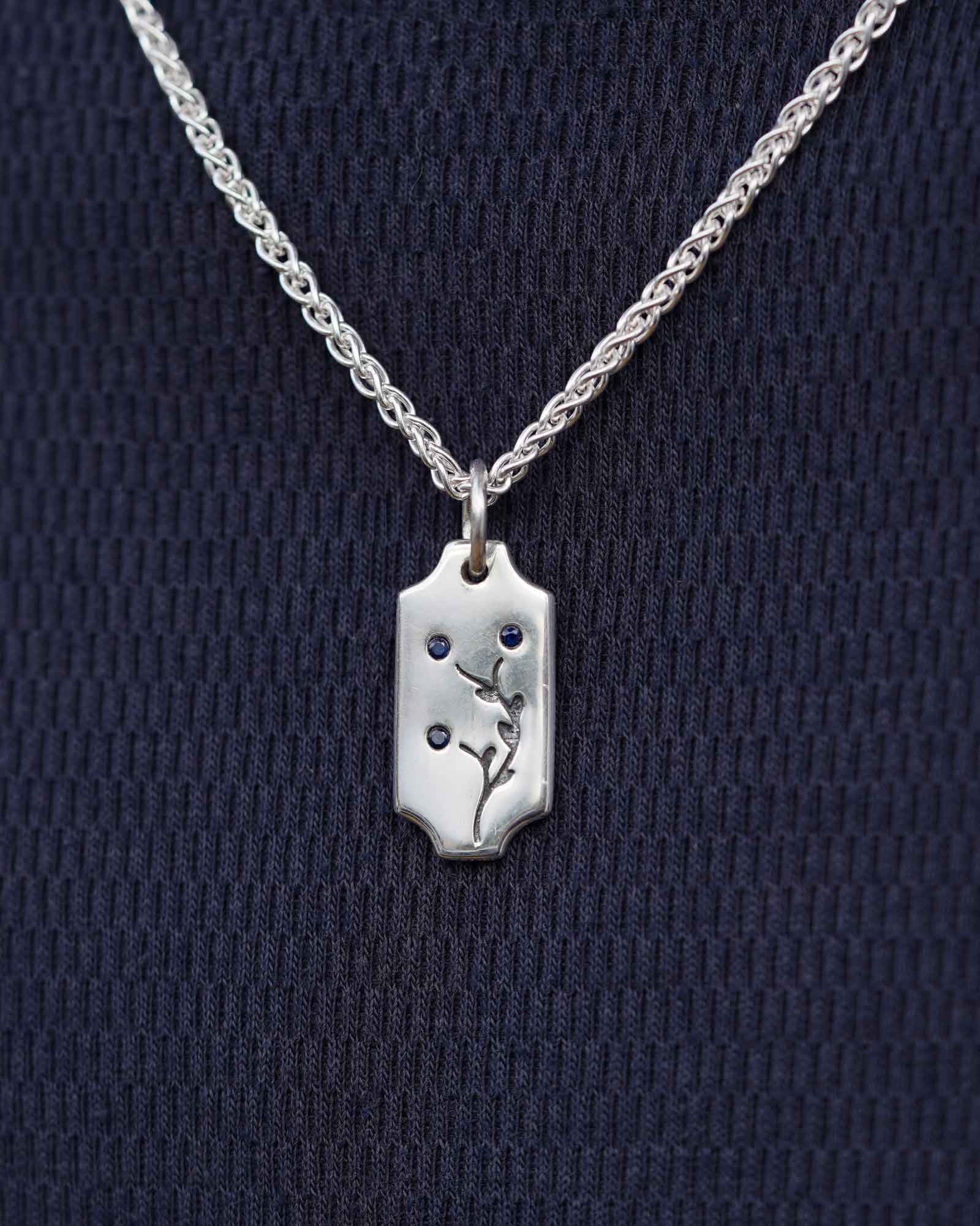 A silver pendant necklace with a small charm featuring a floral design and blue stones, displayed on a textured navy blue background.